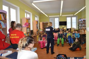 biblioteka miejska w mrągowie, policjantka prowadzi zajęcia dzieci siedzą na krzesełkach, ratownicy wopr stoją z boku