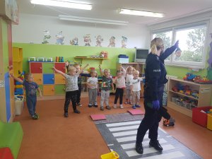 w przedszkolu policjantka stoi przy imitacji pasów leżących na podłodze i pokazuje dzieciom lewą ręką kierunek, dzieci trzymają w górze lewą rękę