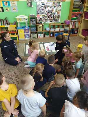 w szkolnej sali na dywanie siedzą dwie policjantki i dzieci. Jedna z policjantek trzyma w ręce książkę i czyta dzieciom bajkę