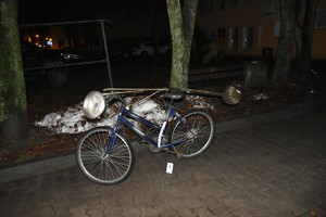 na polbruku przed drzewami stojący rower koloru niebieskiego i przymocowana do niego lampa wolnostojąca