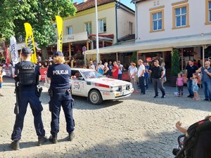 policjant i policjantka stojący na placu, obok nich przejeżdżający samochód rajdowy
