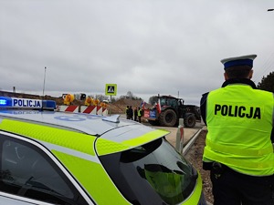 na pierwszym planie stoi radiowóz, obok niego tyłem stoi policjant. W tle widać ciągniki i protestujących rolników